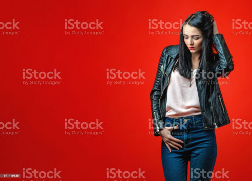 Brooding young girl with black hair in a leather jacket and blue jeans on a bright red background.